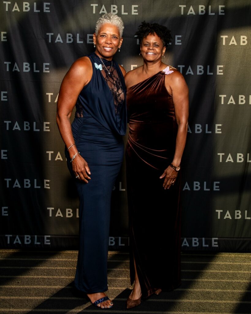 Two women in formal gowns pose for a photo at an event with a "TABLE" backdrop.