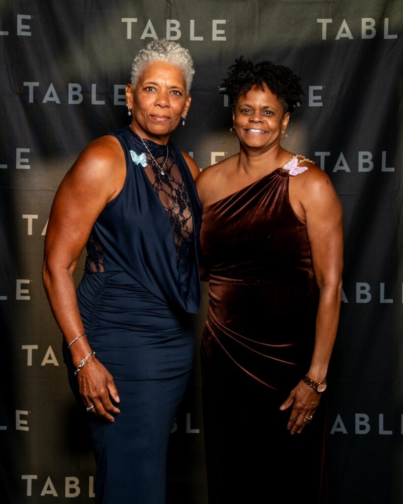 Two women in formal attire pose in front of a backdrop with "TABLE" repeated.