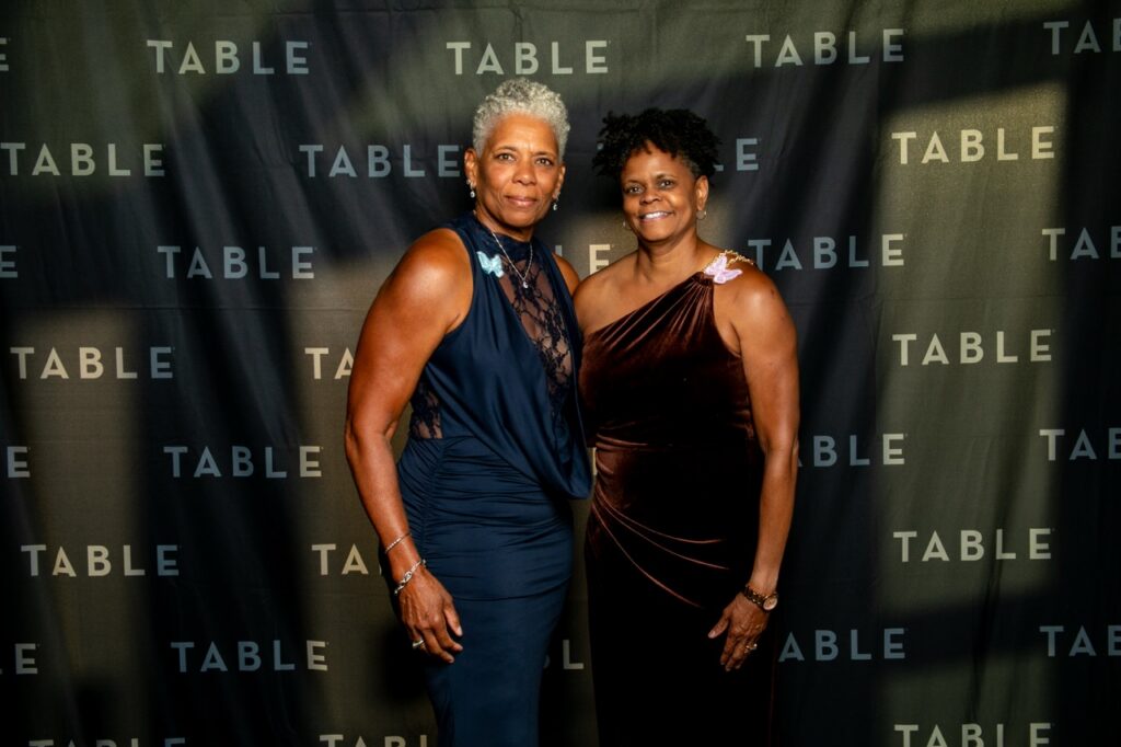 Two women in formal attire pose in front of a backdrop with the word "TABLE" repeated.