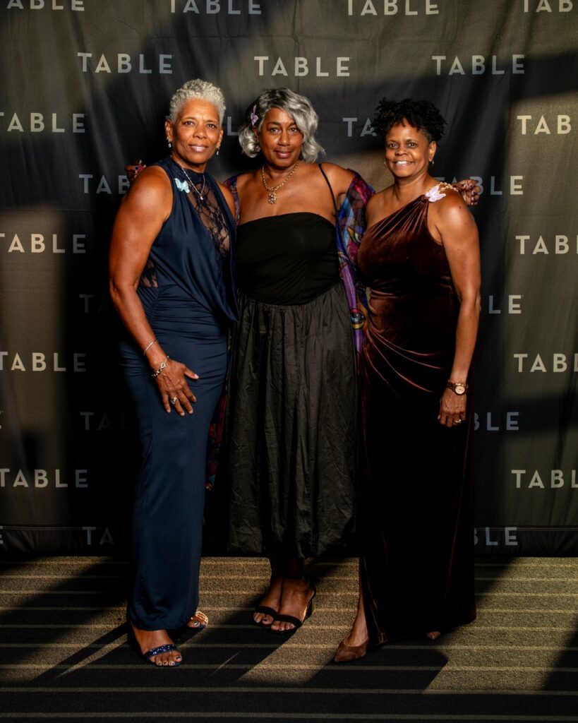 Three elegant women in formal gowns pose for a photo against a black backdrop with "TABLE" logos.