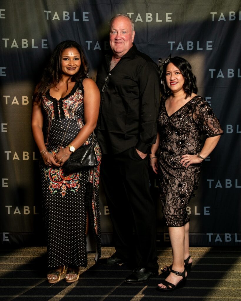 Three people in formal attire pose in front of a backdrop with repeating "TABLE" text.