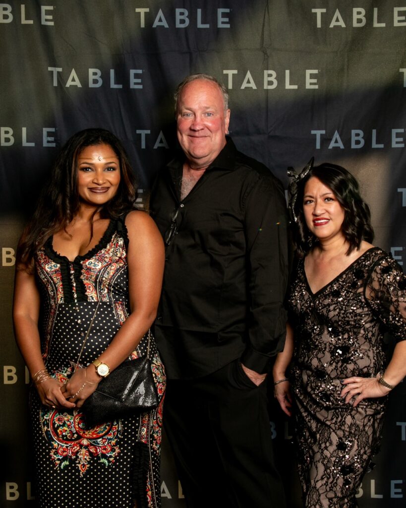 Three people pose for a photo in front of a backdrop with the word "TABLE" repeated.