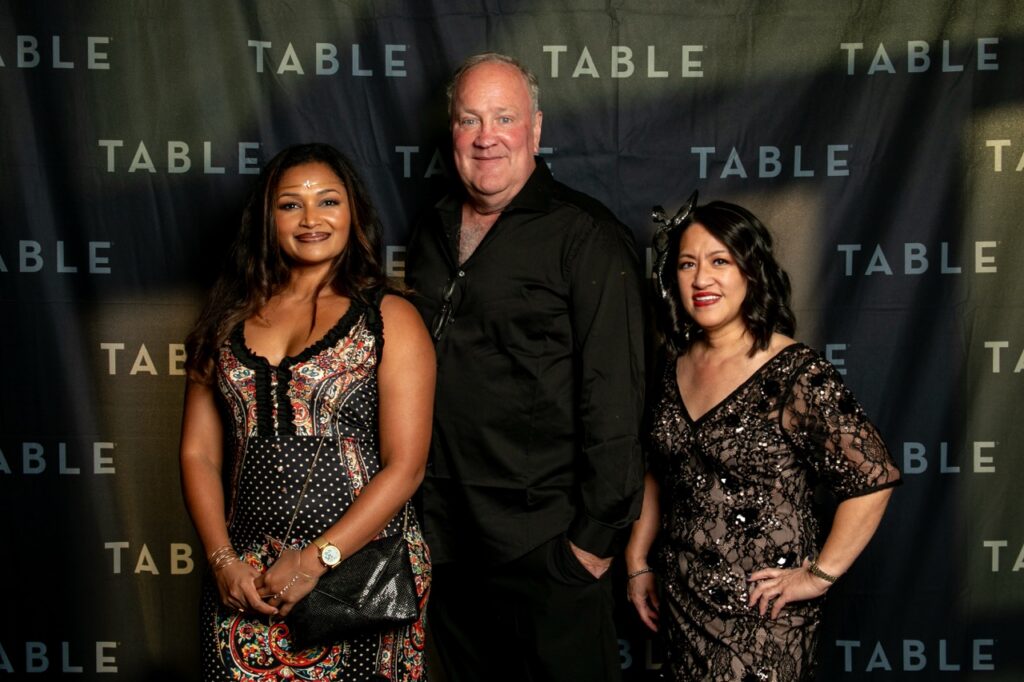 Three people pose for a photo in front of a backdrop with "TABLE" repeated.