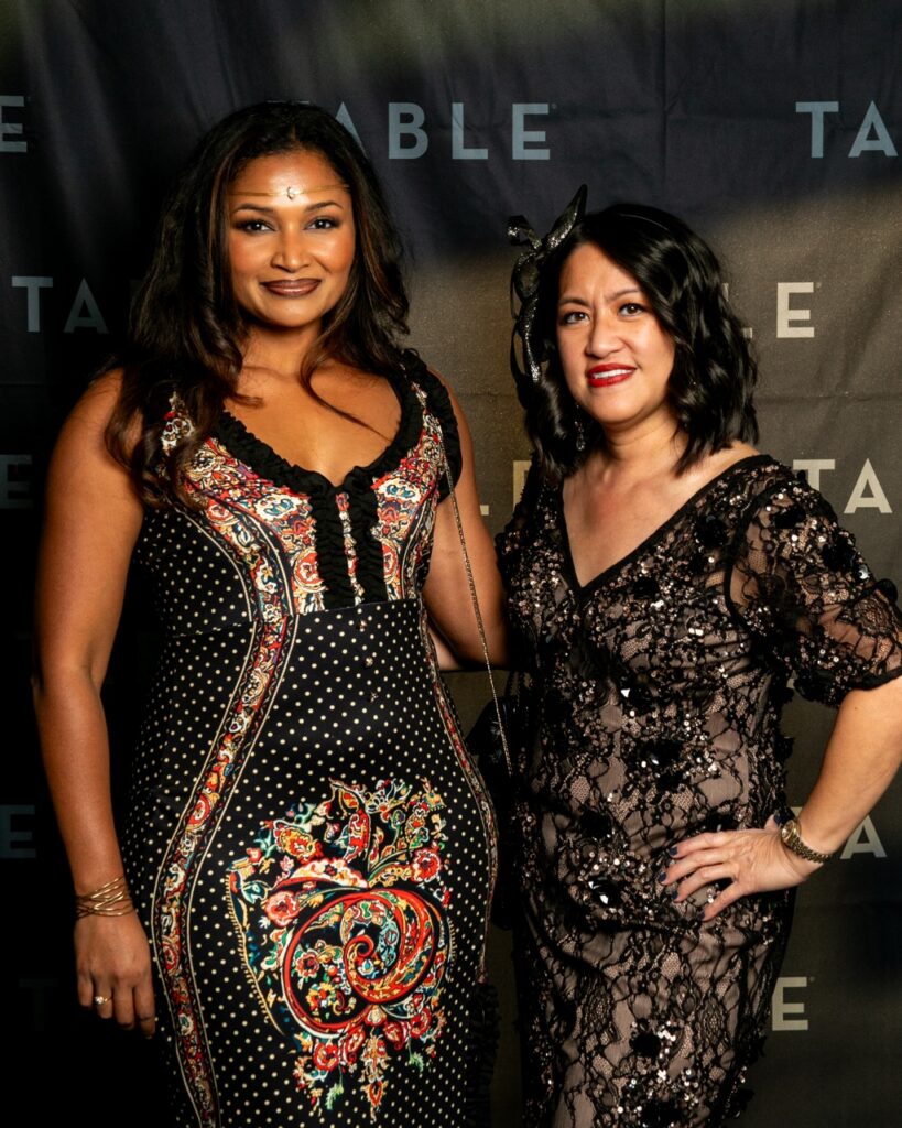Two women pose for a photo in front of a dark backdrop with the word "TABLE" repeated.