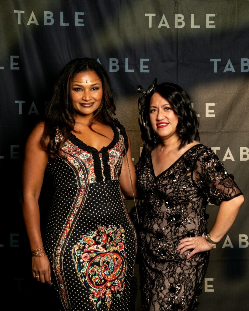 Two women in formal dresses pose in front of a black backdrop with "TABLE" printed repeatedly.