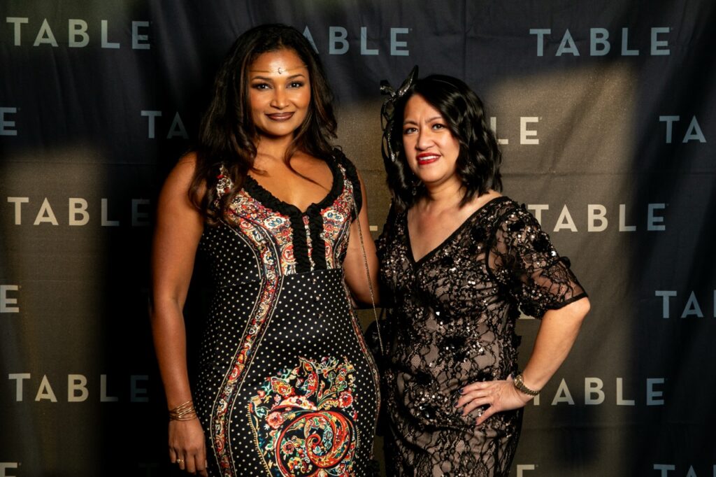 Two women in formal attire pose in front of a backdrop with the word "TABLE" repeated.