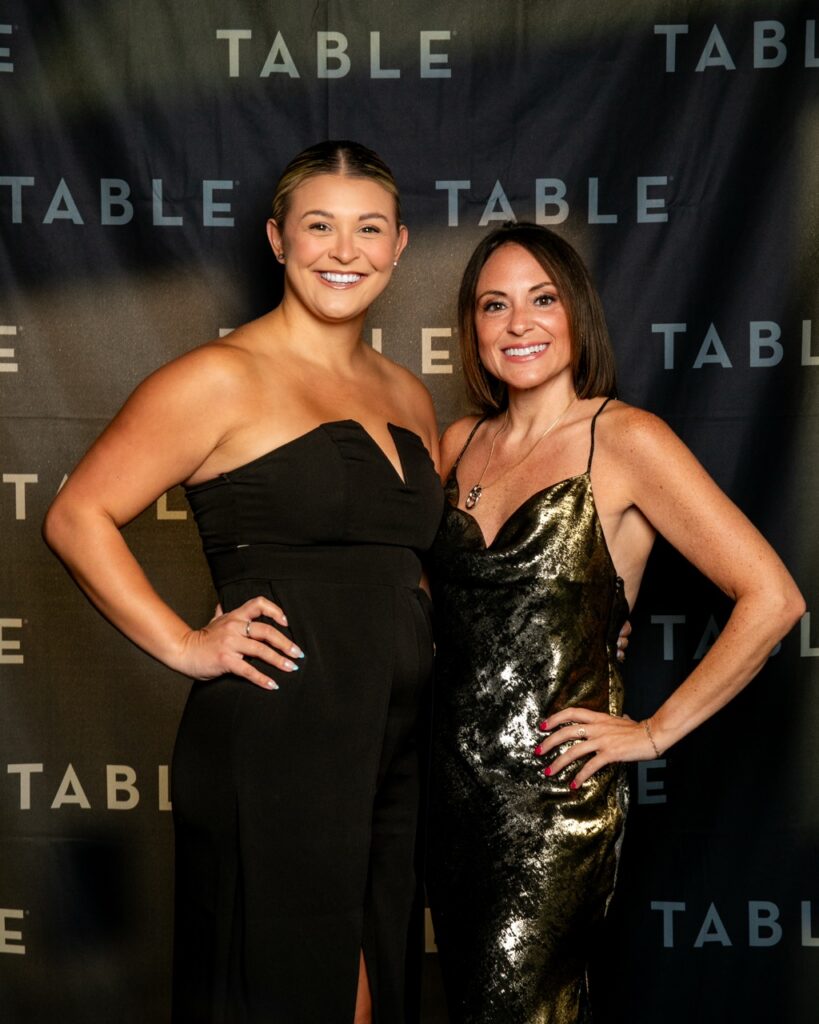 Two women in formal attire posing in front of a black backdrop with the word "TABLE" repeated