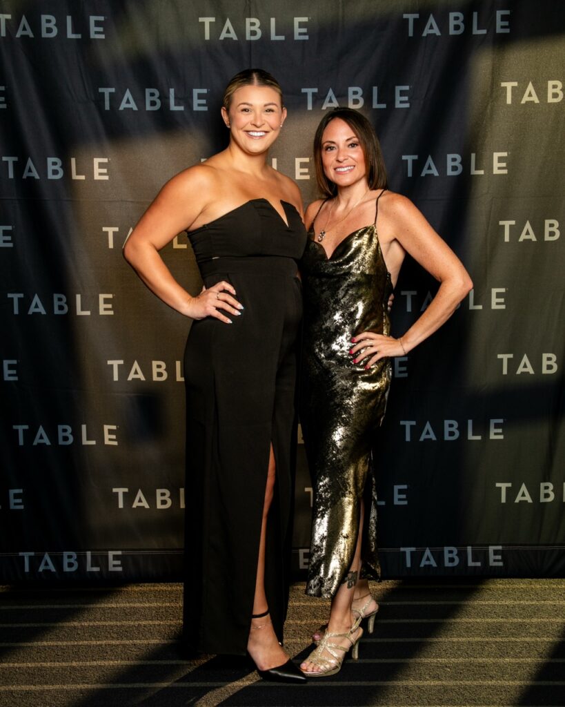 Two women in formal wear pose in front of a "TABLE" backdrop at an event.