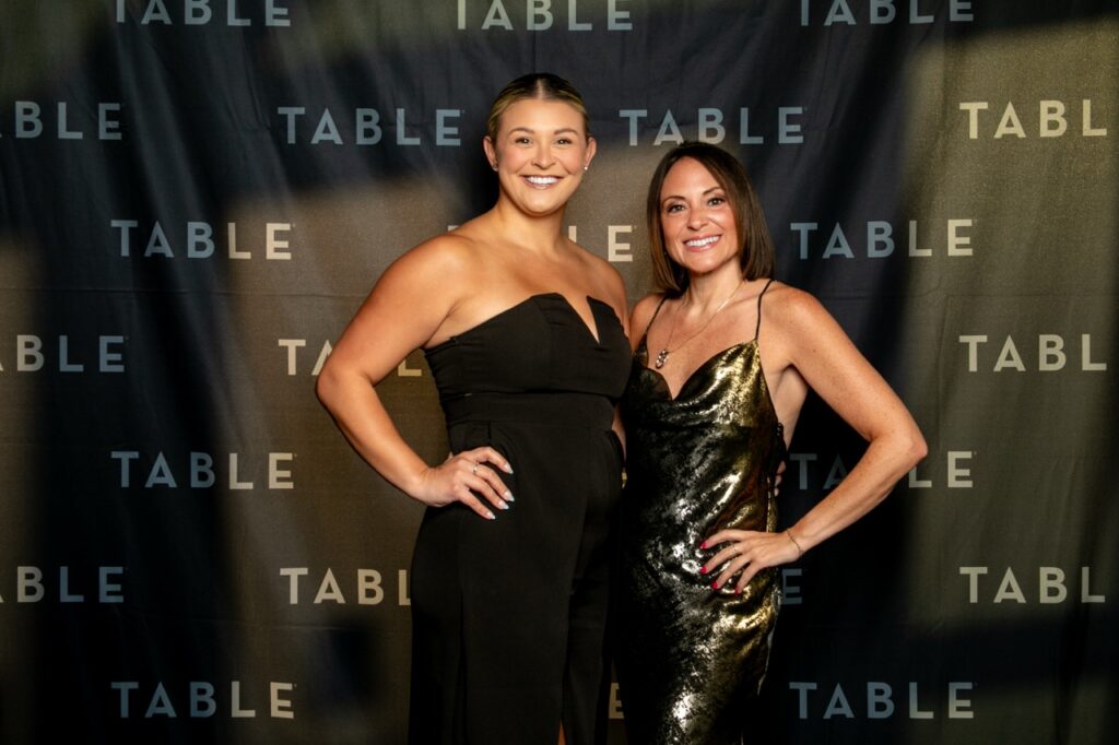 Two women in formal attire pose in front of a backdrop with the word "TABLE" repeated.