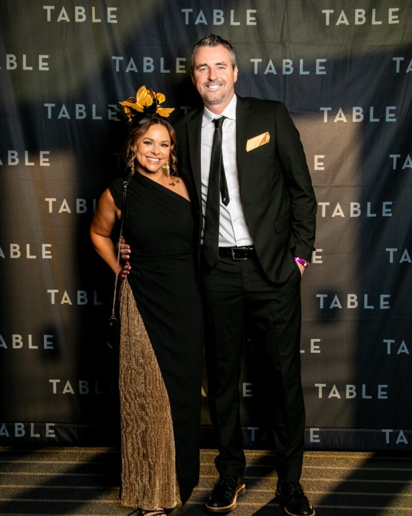 Couple in formal attire at a "TABLE" branded event, woman wears a gold fascinator.