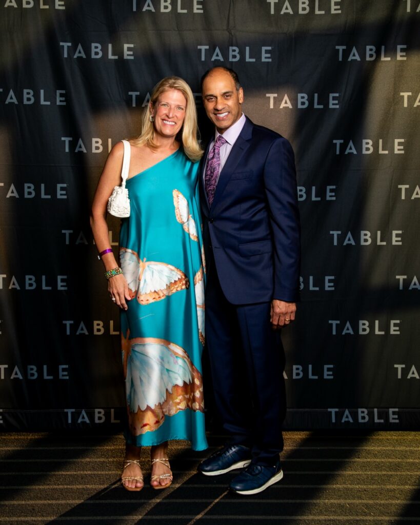 Man and woman pose in front of a TABLE backdrop at an event