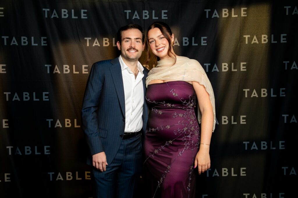 A man in a suit and a woman in a purple dress pose for a photo at an event with a "TABLE" backdrop.