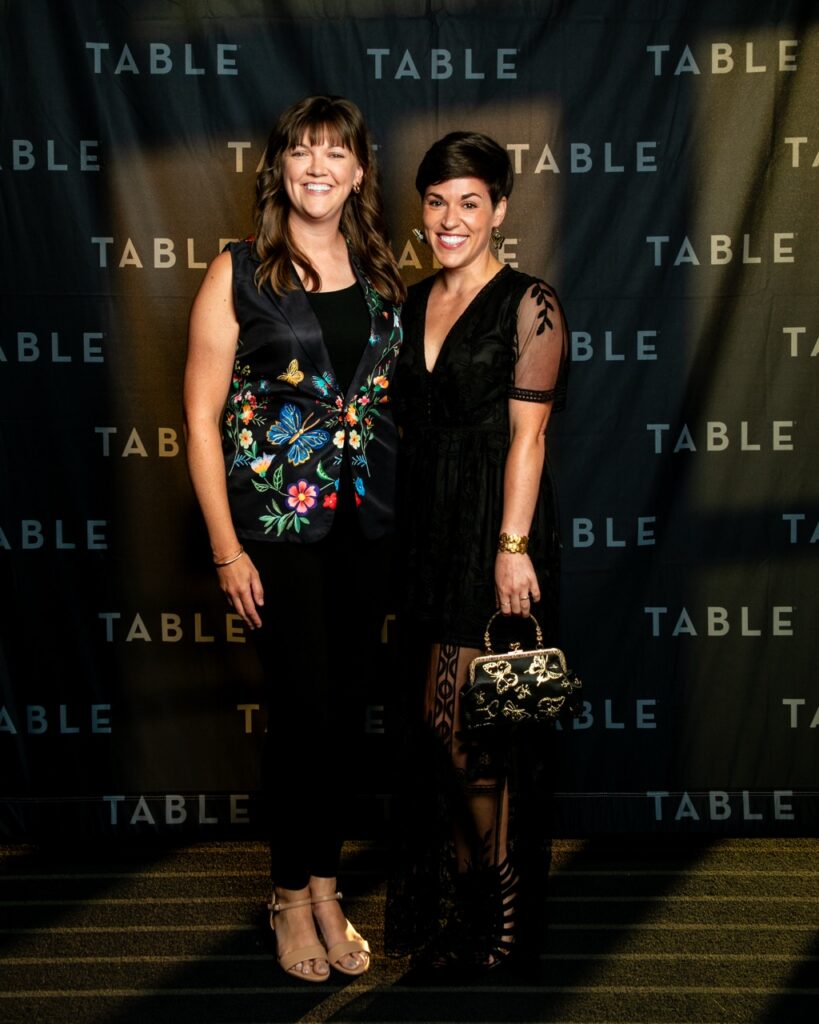 Two women in black dresses pose in front of a backdrop with the word "TABLE" repeated.