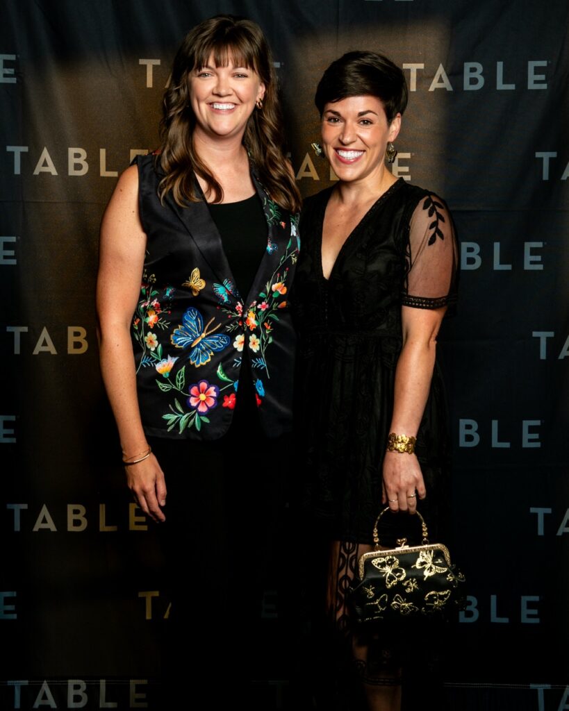 Two women in black outfits smile in front of a dark backdrop with "TABLE" branding.