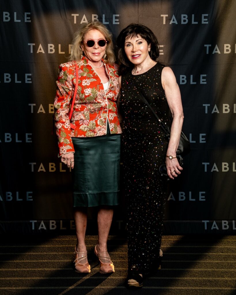 Two women in formal attire pose in front of a backdrop with "TABLE" repeated.