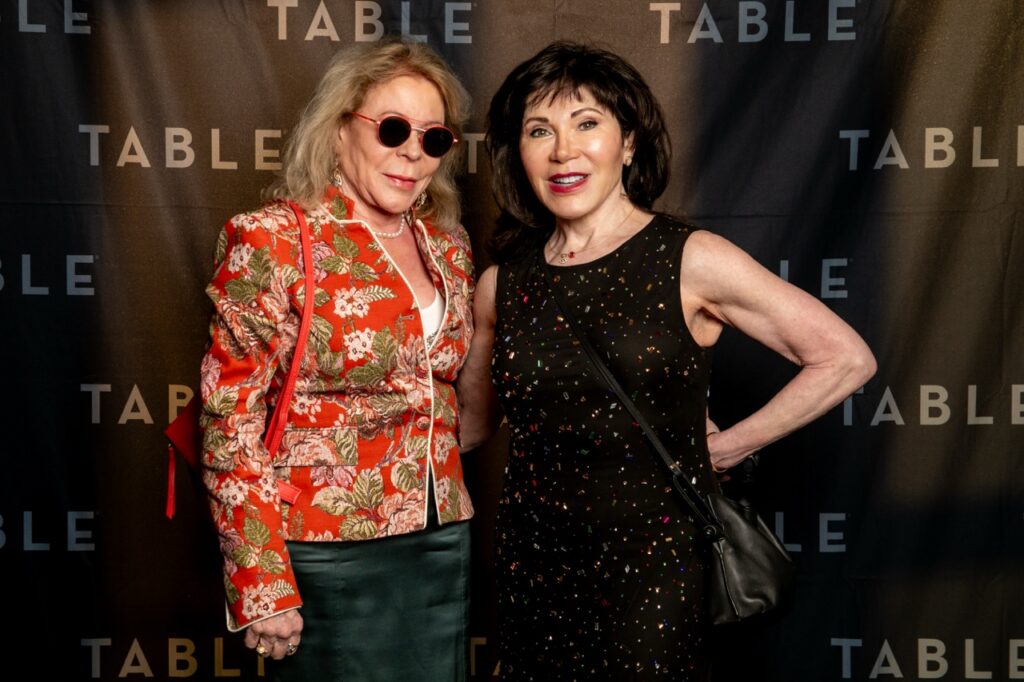 Two women in formal attire pose in front of a backdrop with the word "TABLE" repeated.