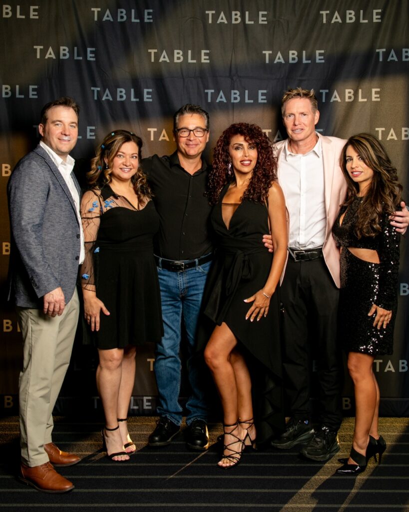 Six people posing in front of a black backdrop with "TABLE" text