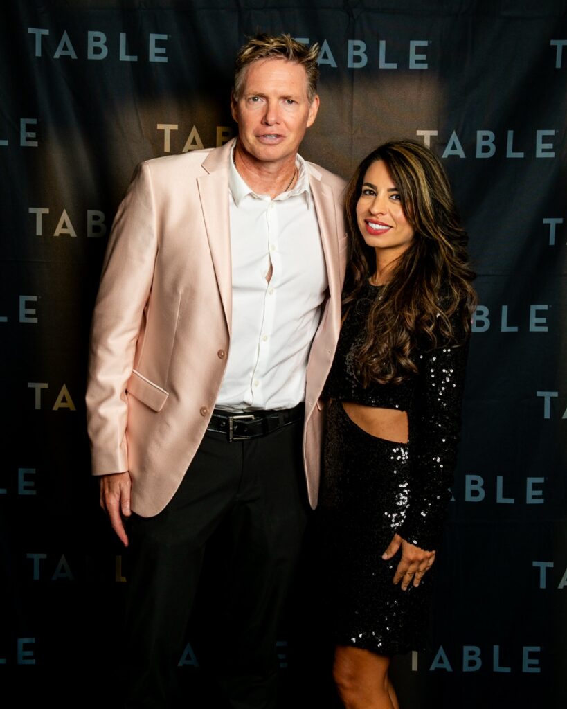 Couple posing for a photo at an event with a repeating "TABLE" backdrop.