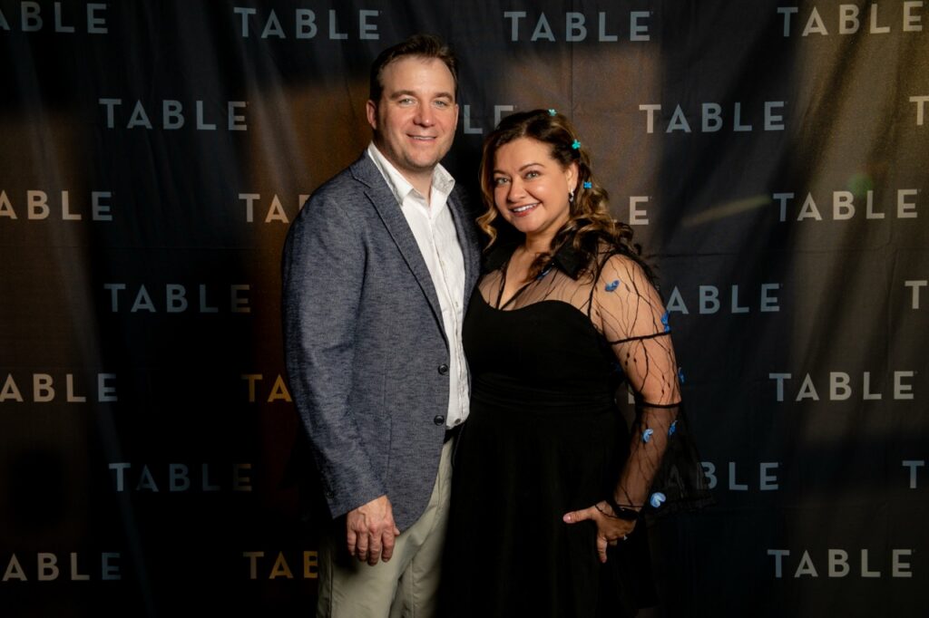 Couple posing for a photo in front of a backdrop with the word "TABLE" repeated