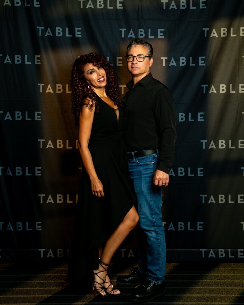 Couple posing in front of a backdrop with "TABLE" repeatedly printed on it.