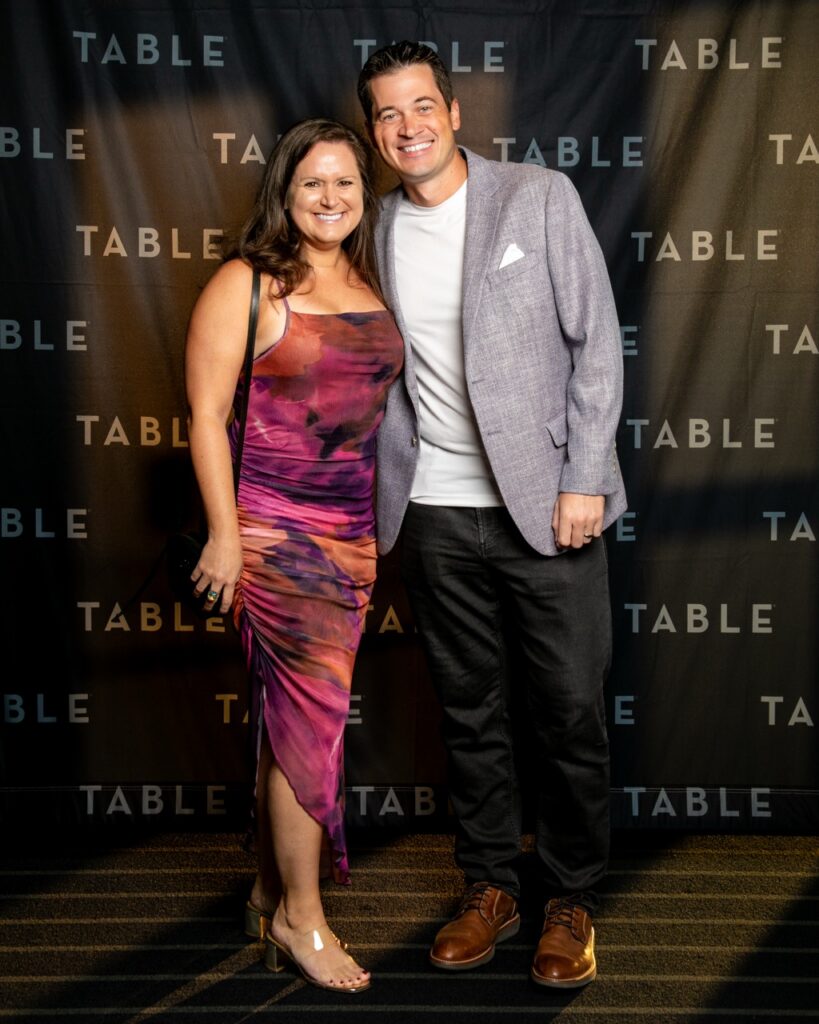 Couple posing in front of a black backdrop with "TABLE" logo at an event.