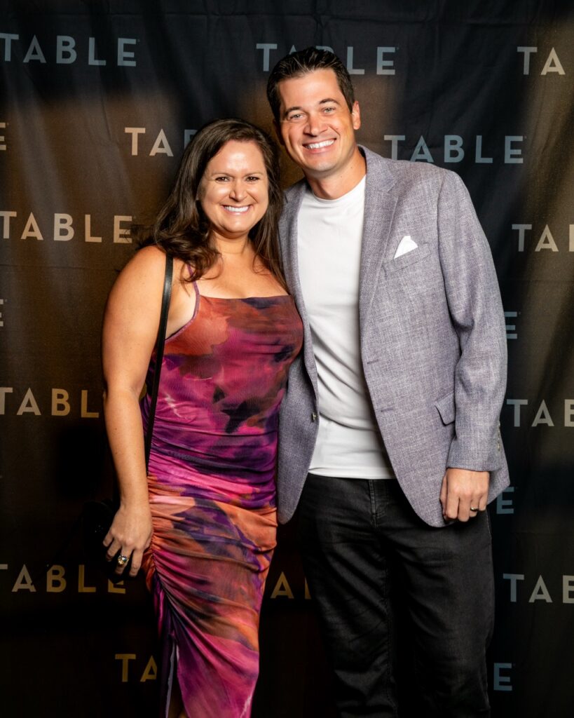 Couple smiling at a TABLE event, woman in a colorful dress, man in a blazer