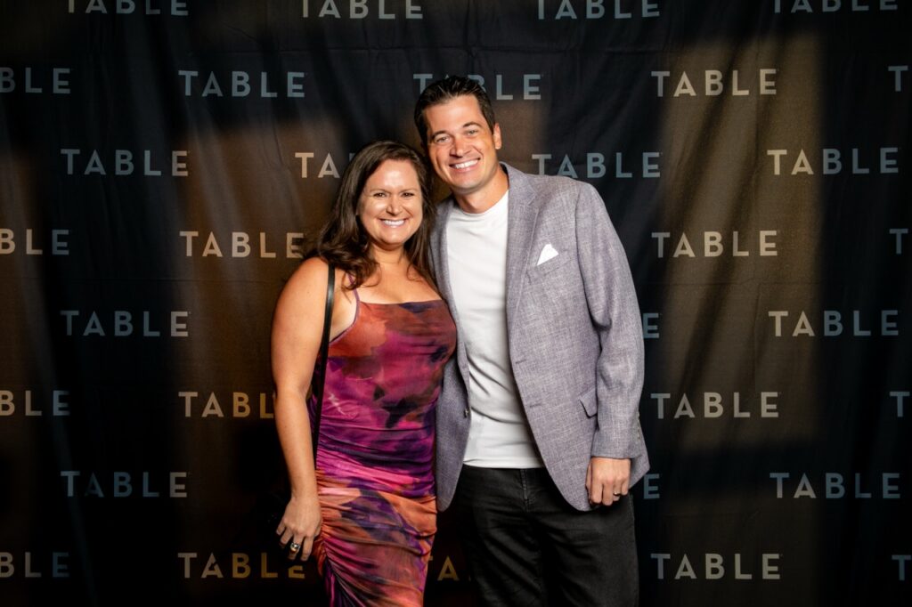 Couple smiles in front of a black backdrop with the word "TABLE" repeated