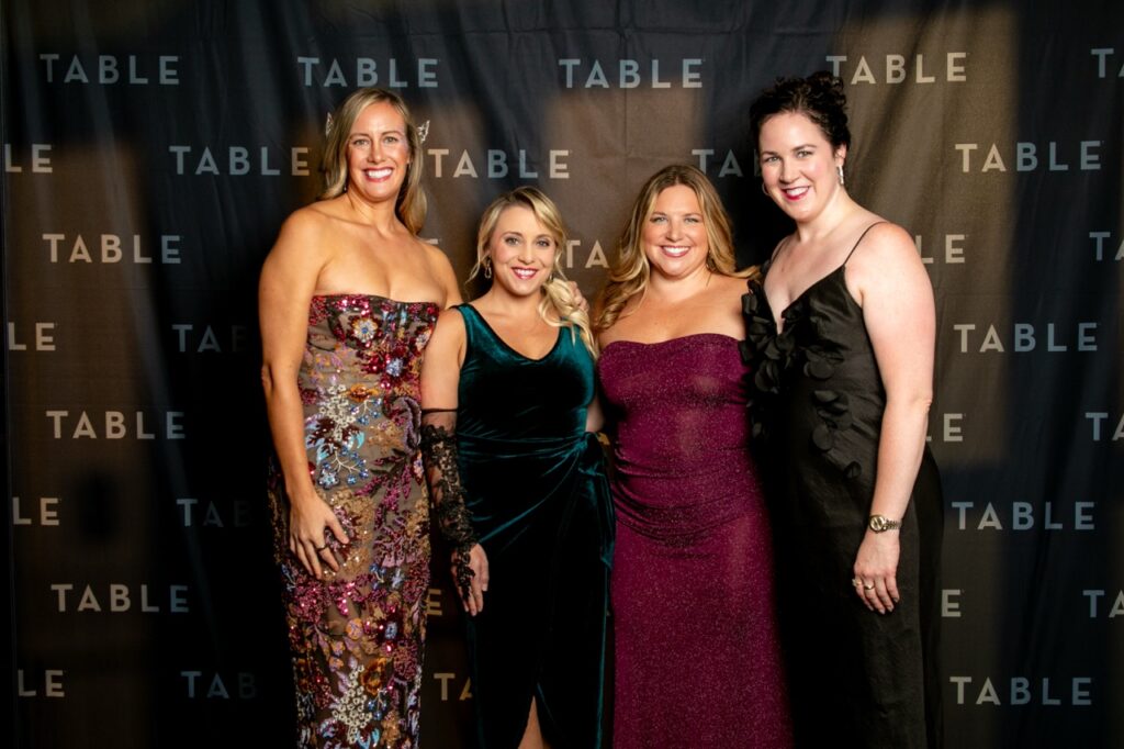 Four women in formal gowns smile in front of a black backdrop with "TABLE" repeated.