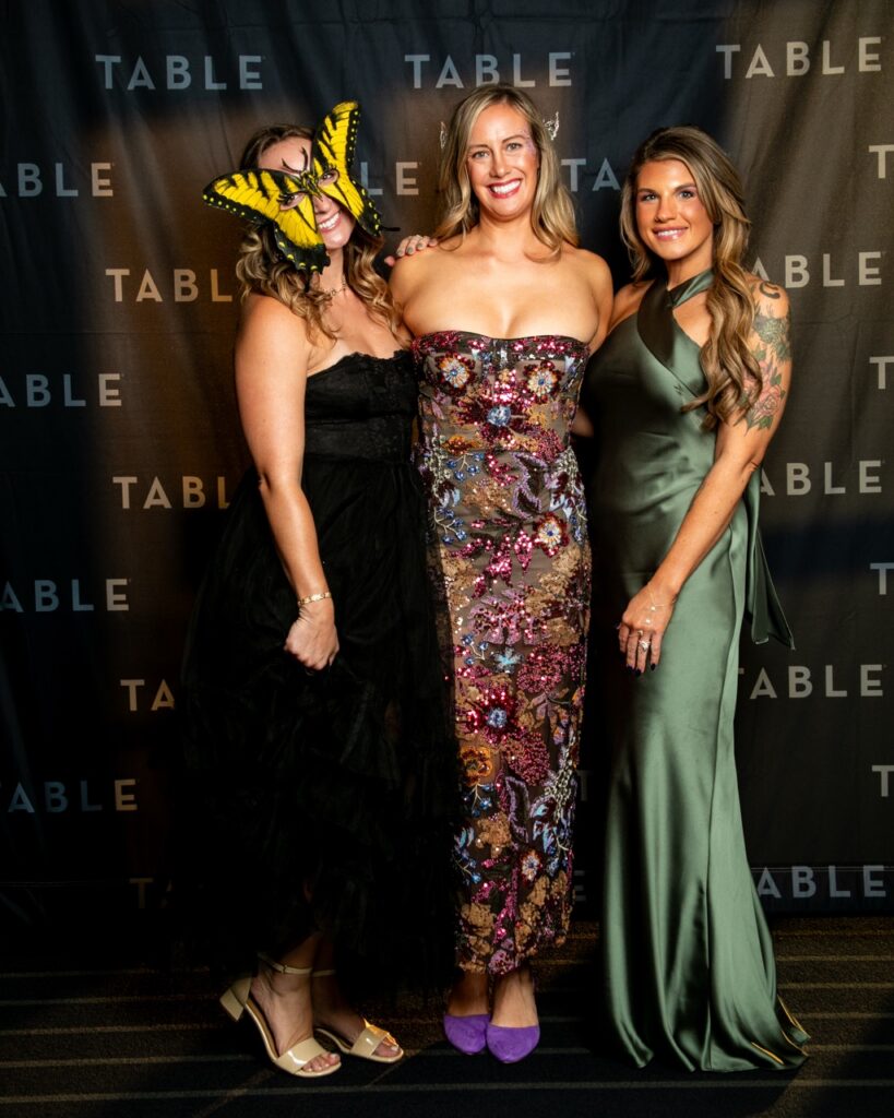 Three women in formal attire pose for a photo at an event with "TABLE" logo backdrop.