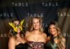 Riverlife’s Party at the Pier 2026 Three women in formal gowns pose for a photo in front of a black backdrop with "TABLE" logos.