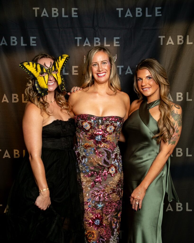 Three women in formal gowns pose for a photo in front of a black backdrop with "TABLE" logos.