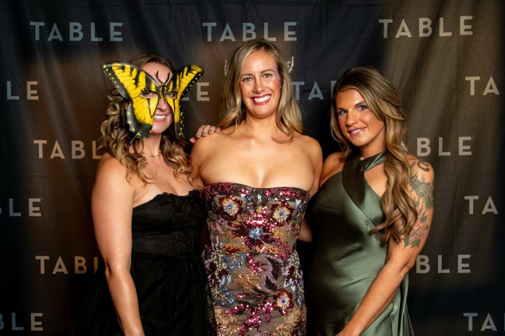 Three women in formal attire pose for a photo at an event with a "TABLE" backdrop.
