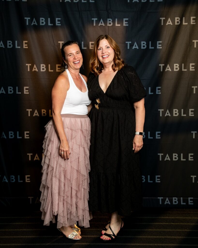 Two women smile in front of a black backdrop with "TABLE" logo.