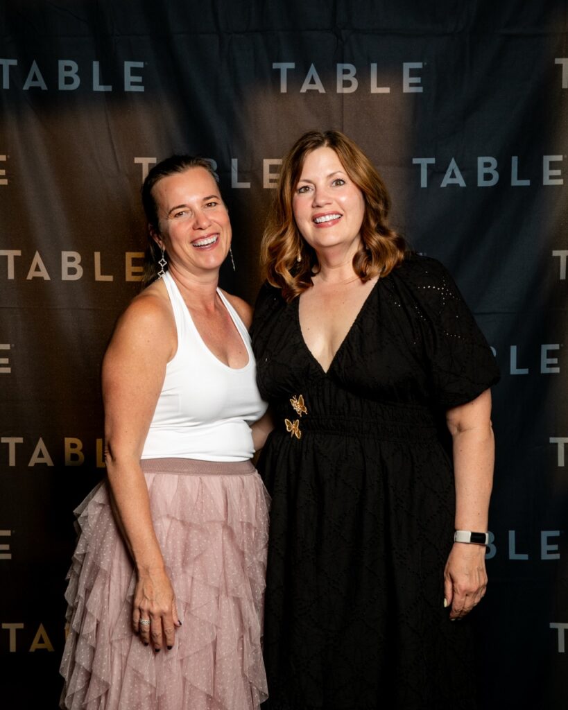 Two women smiling at a TABLE event, one in a white top and pink skirt, the other in a black dress.