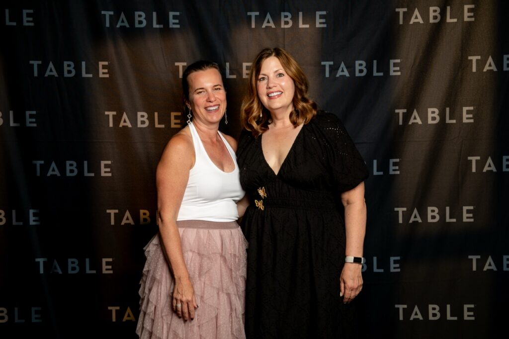 Two women smiling in front of a black backdrop with the word "TABLE" repeated.