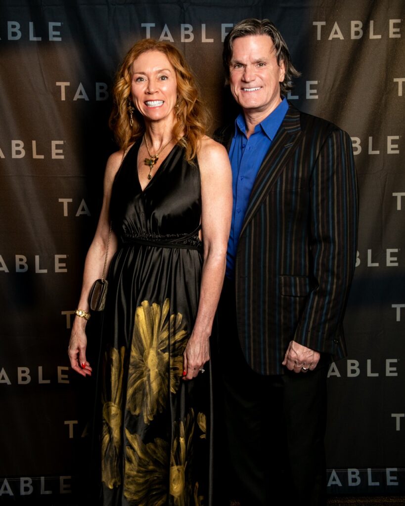 A smiling couple poses for a photo in front of a backdrop with the word "TABLE" repeated.