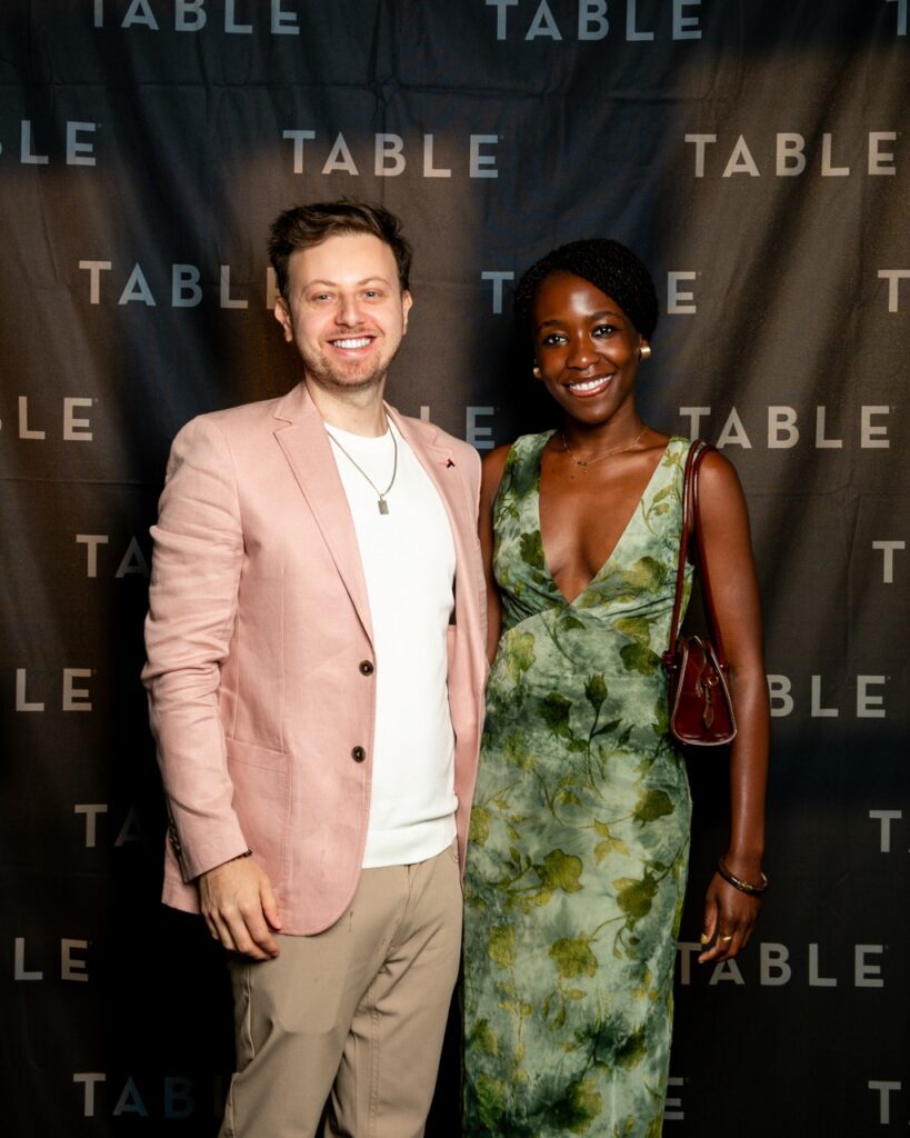 Man in pink blazer and woman in green dress pose in front of TABLE backdrop.