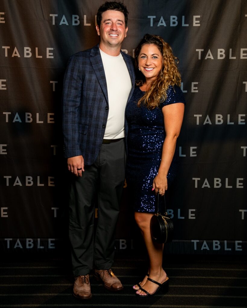 Couple in formal attire poses in front of a TABLE backdrop.