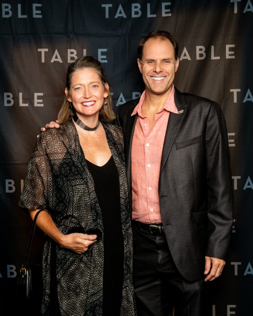 Couple smiles at the camera in front of a TABLE backdrop