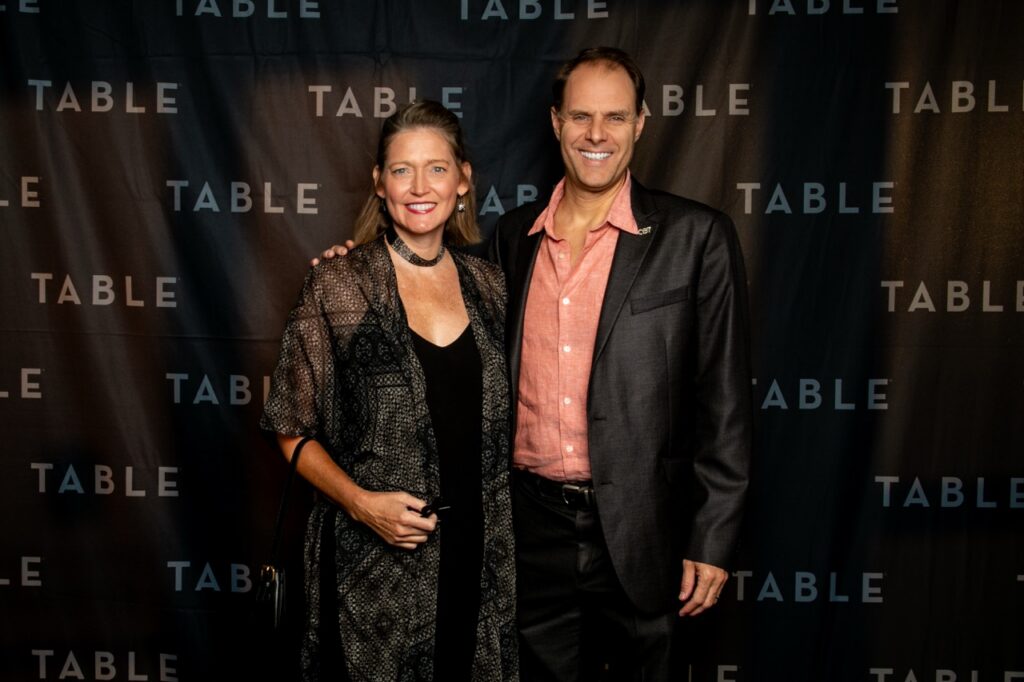 Couple posing in front of a black backdrop with "TABLE" repeated