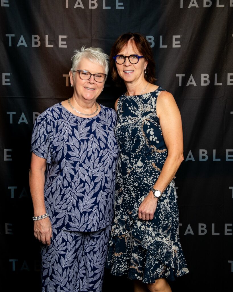 Two women in patterned dresses pose in front of a black backdrop with "TABLE" repeated.