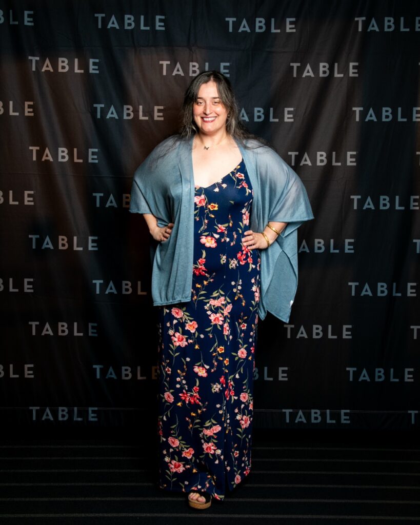 Woman in floral dress and blue shawl poses in front of a "TABLE" backdrop.