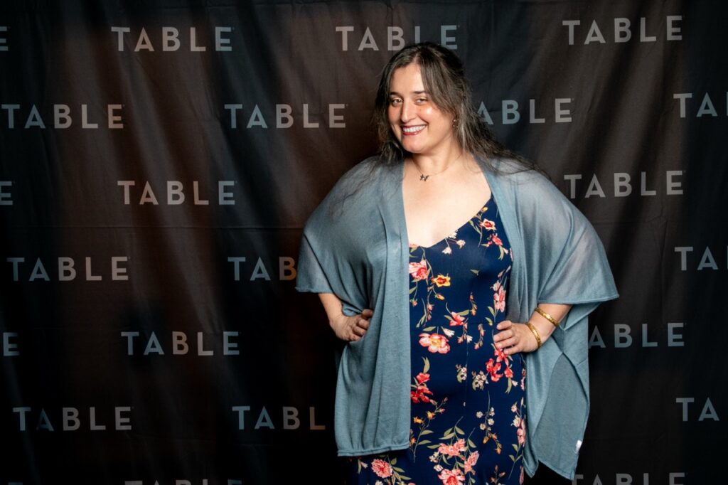 Woman in floral dress and blue shawl poses in front of a "TABLE" backdrop.