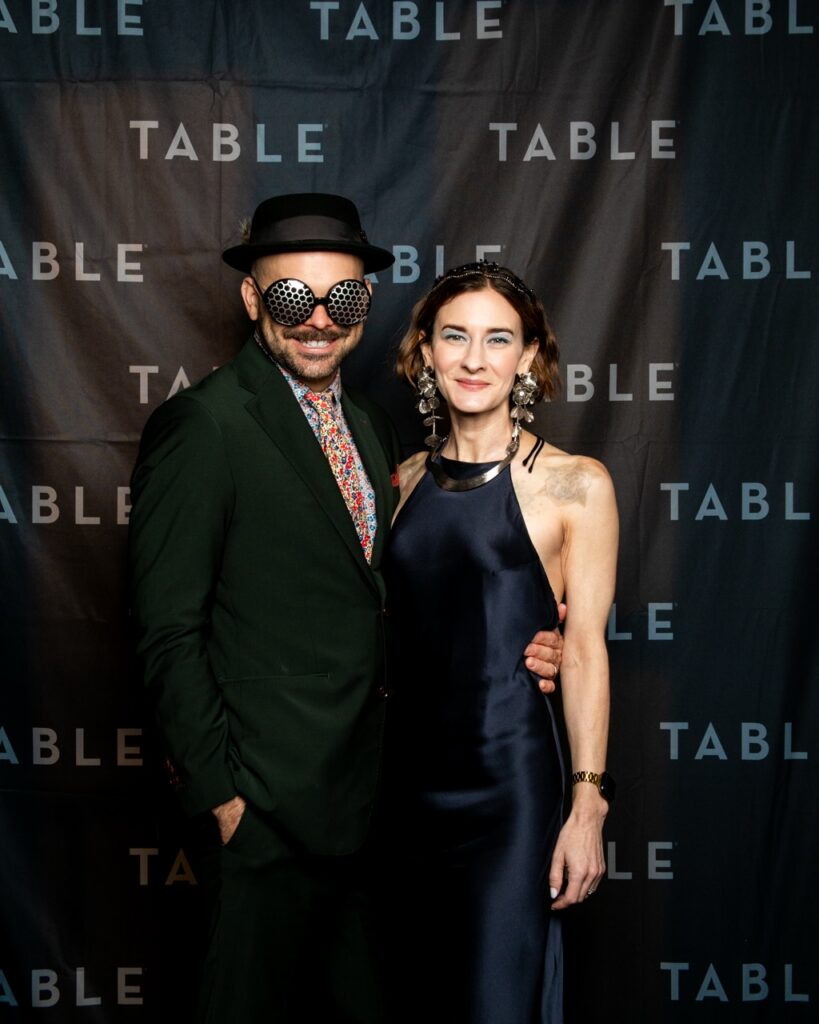 Man in a green suit and hat with goggles, and woman in a navy dress at a TABLE event