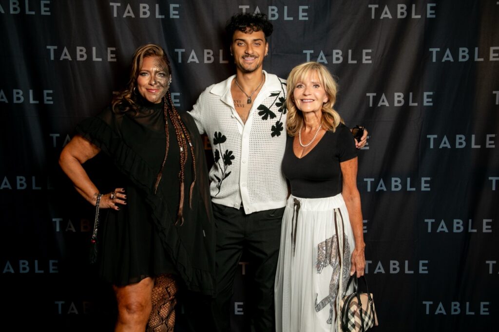 Three people pose for a photo in front of a backdrop with "TABLE" printed repeatedly.