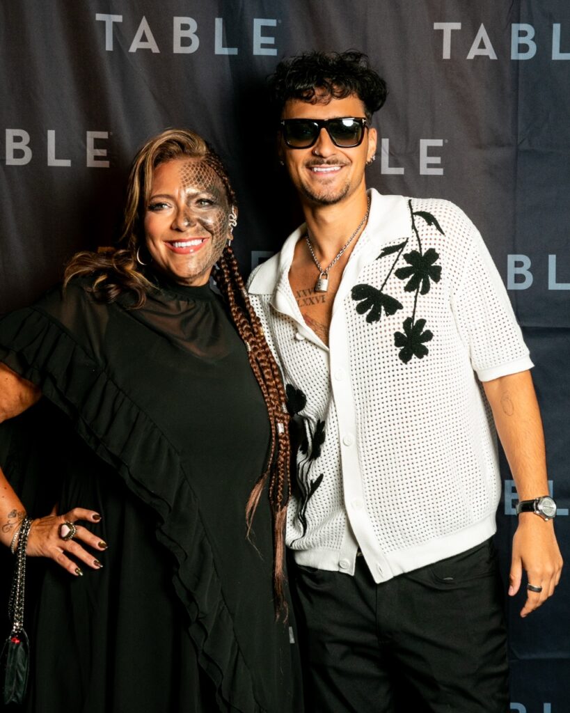 Two people pose in front of a black backdrop with "TABLE" text.