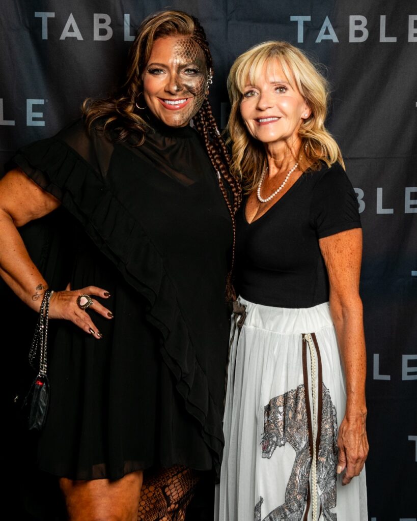 Two women in formal attire pose for a photo at an event with "TABLE" branding.