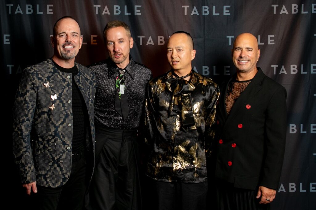 Four men in stylish outfits pose for a photo in front of a backdrop with the word "TABLE" repeated.