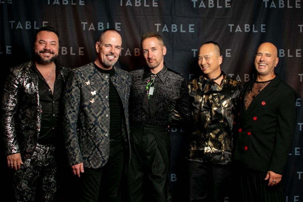 Five men in stylish outfits pose for a photo in front of a backdrop with the word "TABLE" repeated.