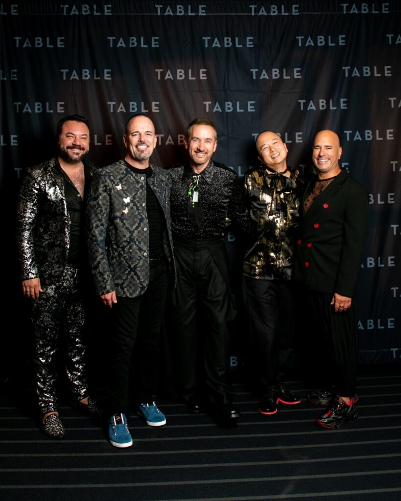 Five stylish men in formal wear pose for a photo at an event.